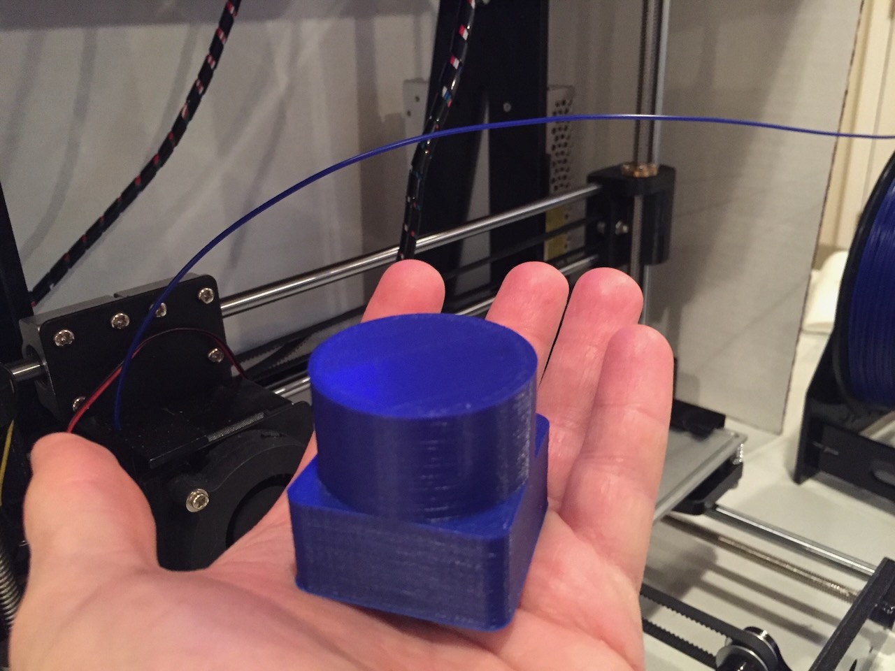 a hand holding a very simple 3D print (a cylinder on a cube) in the background is a 3D printer